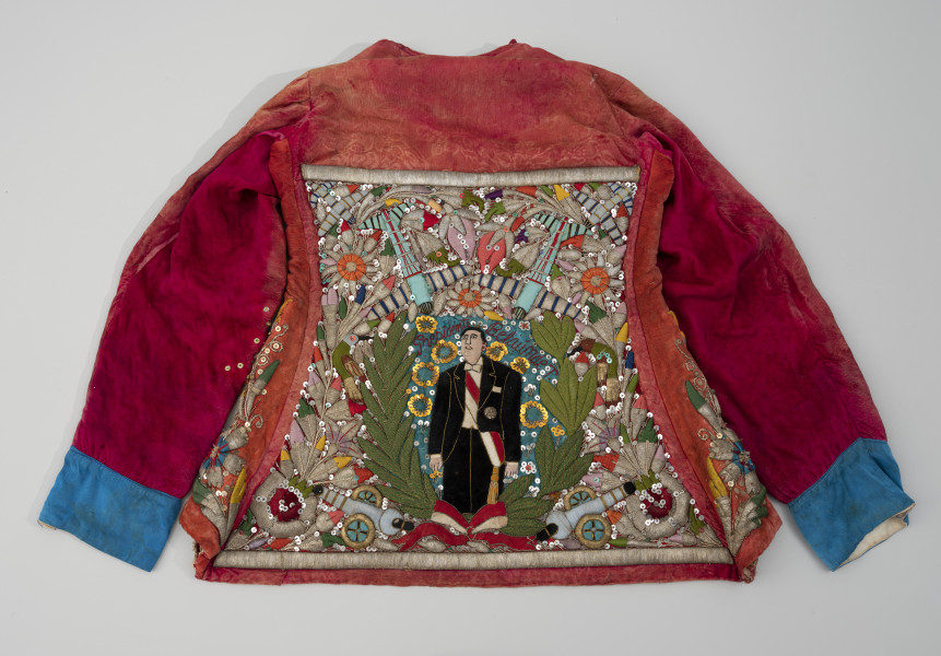 kaftan do tańca negritos - Ujęcie z tyłu; Czerwony, lekko wyblakły, welurowy kaftan o trapezowatym kształcie i luźnym kroju, bogato haftowany, ozdobiony kolorowymi cekinami i szklanymi paciorkami. Tył usztywniony. Na niebieskim tle naszyto postać ubranego w galowy strój peruwiańskiego prezydenta - Fernando Belaúnde Terrego  w otoczeniu armat, samolotów, ptaków (kolibrów?) i kwiatów. Postać w ujęciu do kolan, otoczona zielonym wieńcem przewiązanym czerwono-białą wstęgą. Nad postacią, czerwoną nicią, naszyto napis. Rękawy wykończono niebieską satyną, na welurowej tkaninie zauważalne odbijające się wzory kwiatowe. Na tylnej części rękawów widoczny kolor fuksji. Na czerwonym welurze miejscami widoczne pozostałości odciskanego wzoru kwiatowego. Widoczne naprawy - ślady szycia.