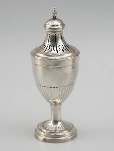 cukiernica do cukru pudru - Ujęcie z tyłu; Puszka z ażurową pokrywą, kształtem nawiązująca do antycznych urn. Wsparta na okrągłej stopie z trzema uskokami, przechodzącej w niewysoki, drobno kanelowany trzon. Korpus smukły, o kielichowej formie, w dolnej części również kanelowany. Pokrywa wysoka, o wklęsłych ściankach zakończona półkolistą kopułką, ze zwieńczeniem w formie maleńkiej wazy. Wycięcia na pokrywie układają się we wzór stylizowanych kampanul. Na cokoliku stopy w prostokątnym polu o zaokrąglonych narożnikach napis VOG | LAER