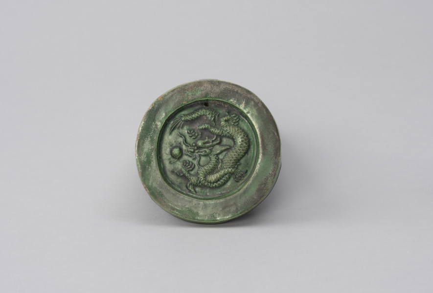 gąsior początkowy ozdobny - Ujęcie z góry; Półcylindryczna dachówka typu gąsior z zewnątrz szkliwiona zieloną glazurą. Zasklepiona okrągłą, ozdobą plakietką z odciśniętym, reliefowym wizerunkiem smoka. Smok zieje ogniem, ma rogi, długie wąsy, grzywę, cztery łapy, wijące się, długie, łuskowate ciało. Obok jego paszczy widać płonącą perłę. W plakietce, nad smokiem znajduje się mały, okrągły otwór. Na dachówce widoczne przetarcia, rysy, ubytki, spękania oraz odbarwienia.