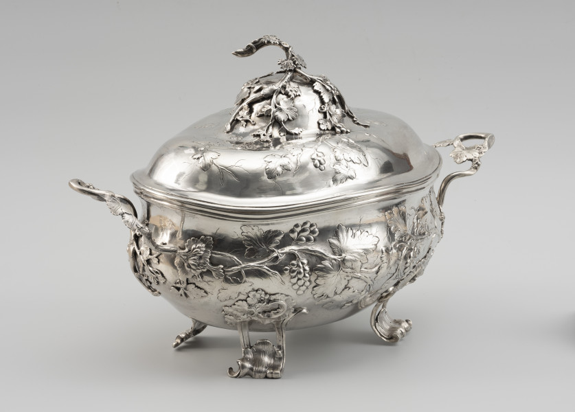 waza - Ujęcie ze skosu z lewej; Owalna teryna, wsparta na czterech nóżkach. Korpus o płynnym, organicznym kształcie i falistej linii brzegu, w dolnej części wybrzuszony. Pokrywa wysoka, dwustopniowo wybrzuszona. Zarówno korpus, jak i pokrywa zdobione repusowanymi w płytkim reliefie naturalistycznymi gałązkami winorośli z gronami owoców. Reliefowy ornament miękko przechodzi w wykonane techniką odlewu, plastyczne gałązki winnej latorośli, tworzące dwa uchwyty przy korpusie oraz ozdobne zwieńczenie pokrywy. Nóżki wolutowo wygięte, ażurowe, zdobione rocaillami i gałązkami winorośli.