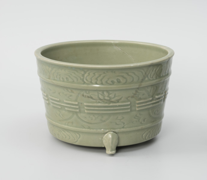 donica ozdobna - Ujęcie z przodu; Kamionkowa donica z trzema dekoracyjnymi nóżkami, widoczna jedna, szkliwiona celadonem w jasnozielonym odcieniu. Ma kształt odwróconego, ściętego stożka. Jej nogi stylizowane są na łapy zwierzęce, zostały doklejone do wykonanego na kole garncarskim korpusu. Powierzchnia naczynia została ozdobiona wklęsło-wypukłym ornamentem reliefowym, podzielonym na trzy poziome strefy oddzielone dookolnie biegnącymi listwami. Górny i dolny pas wypełniono motywami roślinnymi – najprawdopodobniej peoniami, środkowa część natomiast składa się ze wzoru roślinnego (peonie) oraz wypukłych linii ułożonych w różnych sekwencjach, przedstawiających osiem podstawowych trygramów. Na ściankach z przodu widoczne ślady klejenia oraz drobne ubytki.