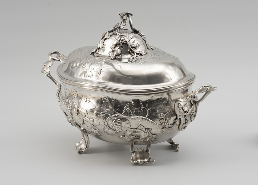 waza - Ujęcie ze skosu z prawej; Owalna teryna, wsparta na czterech nóżkach. Korpus o płynnym, organicznym kształcie i falistej linii brzegu, w dolnej części wybrzuszony. Pokrywa wysoka, dwustopniowo wybrzuszona. Zarówno korpus, jak i pokrywa zdobione repusowanymi w płytkim reliefie naturalistycznymi gałązkami winorośli z gronami owoców. Reliefowy ornament miękko przechodzi w wykonane techniką odlewu, plastyczne gałązki winnej latorośli, tworzące dwa uchwyty przy korpusie oraz ozdobne zwieńczenie pokrywy. Nóżki wolutowo wygięte, ażurowe, zdobione rocaillami i gałązkami winorośli.