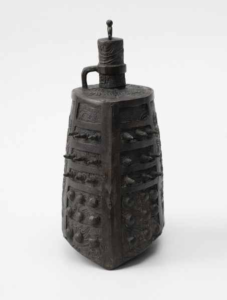 zhong (dzwon chiński) - ujęcie z lewej; Wykonany z brązu dzwon o soczewkowatym przekroju poprzecznym. Na płaskim hełmie znajduje się korona w formie trzpienia, zdobionego rysunkami ptaków, z małym pałąkowatym uszkiem na szczycie zwieńczonym kuleczką i drugim, wygiętym pod kątem prostym, wychodzącym z hełmu i połączonym z trzpieniem za pomocą okrągłej obejmy. Na bogaty ornament reliefowy w postaci siatki wzoru geometrycznego (meander) i zoomorficznego (przedstawienia wężowatych stworzeń, być może smoków) po bokach nałożono kolce mei (w górnej części) oraz guzki (w dolnej części). Dzwon tego typu nie posiada serca, jego środek pozostaje pusty.