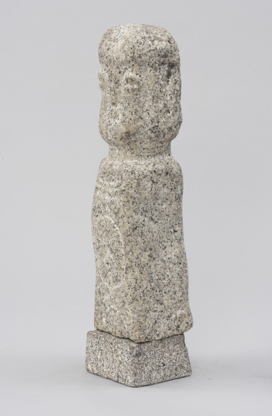 rzeźba; Polelum - ujęcie przodu ze skosu z prawej; Monolityczna, antropomorficzna pionowa forma o cechach ludzkich zredukowanych do prostopadłościennego korpusu i osadzonej na krótkiej szyi głowy. Głowa o miękko zaznaczonych rysach twarzy, wydobytych negatywowym szlifem powierzchni wokół nosa, oczu, ust i policzków, który nakreśla także linię fryzury. Korpus ozdobiony rytem owalu, skierowanej w górę strzały oraz trzech poziomych linii powyżej. Figura ustawiona na nieco szerszym, kamiennym postumencie.