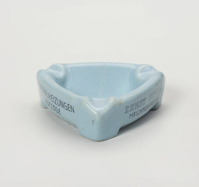 popielniczka reklamowa - Ujęcie z boku; Popielniczka ceramiczna w kolorze szaroniebieskim o kształcie trójkąta równoramiennego z zaokrąglonymi narożnikami. Wywinięty na zewnątrz kołnierz tworzy boki naczynia, wydłużone na narożnikach w formę nóżek-podstawek. W górnej wypłaszczonej części kołnierza znajdują się zagłębienia służące do odłożenia papierosa. Na dwóch bocznych ściankach napisy reklamowe w 2 wierszach ciemnoniebieskimi literami.
