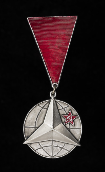 medal - Ujęcie z przodu; Odznaczenie w formie medalu na trójkątnej zawieszce. Medal przybiera kształt koła symbolizującego planetę Ziemię, z naniesionymi liniami południków i równoleżników, na której umieszczono radziecką gwiazdę z sierpem i młotem oraz wypukłą gwiazdę trójramienną, której wierzchołki wychodzą poza obręb koła. Zawieszka ma formę odwróconego wierzchołkiem do dołu trójkąta równoramiennego o podniesionych krawędziach i wypełniona jest czerwoną emalią o prążkowanej strukturze. Zawieszka połączona z medalem za pomocą kółka mosiężnego.