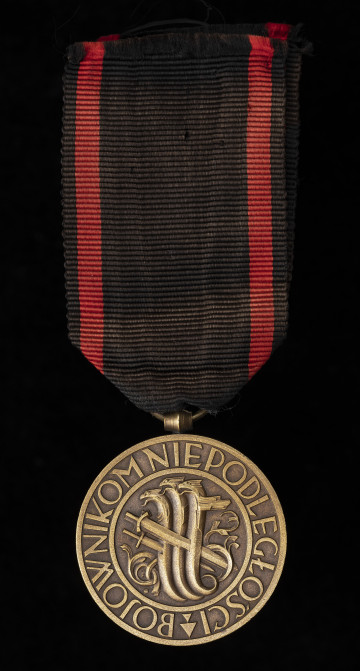 MNS/H/635 Medal Niepodległości - Ujęcie z przodu; Medal w kształcie okręgu, na którego awersie, w polu znajduje się wyobrażenie 3 hydr, przebitych trzema mieczami. Całość zamknięta w podwójnym liniowym otoku zawierającym napis BOJOWNIKOM NIEPODLEGŁOŚCI. Zawieszka z przylutowanego uszka o kształcie beczułkowatym i przewleczonego, zlutowanego kółeczka. Wstążka czarna z dwoma czerwonymi, pionowymi pasami.