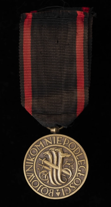 MNS/H/635 Medal Niepodległości - Ujęcie z przodu; Medal w kształcie okręgu, na którego awersie, w polu znajduje się wyobrażenie 3 hydr, przebitych trzema mieczami. Całość zamknięta w podwójnym liniowym otoku zawierającym napis BOJOWNIKOM NIEPODLEGŁOŚCI. Zawieszka z przylutowanego uszka o kształcie beczułkowatym i przewleczonego, zlutowanego kółeczka. Wstążka czarna z dwoma czerwonymi, pionowymi pasami.