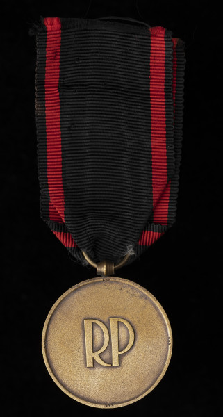 Medal Niepodległości - Ujęcie z tyłu; Medal w kształcie okręgu, na którego rewersie, pośrodku litery RP w polu zamkniętym pojedynczym otokiem z czterema grupami potrójnych nacięć na krawędzi w odstępach 90º. Zawieszka z przylutowanego uszka o kształcie beczułkowatym i przewleczonego, zlutowanego kółeczka. Wstążka czarna z dwoma czerwonymi, pionowymi pasami.