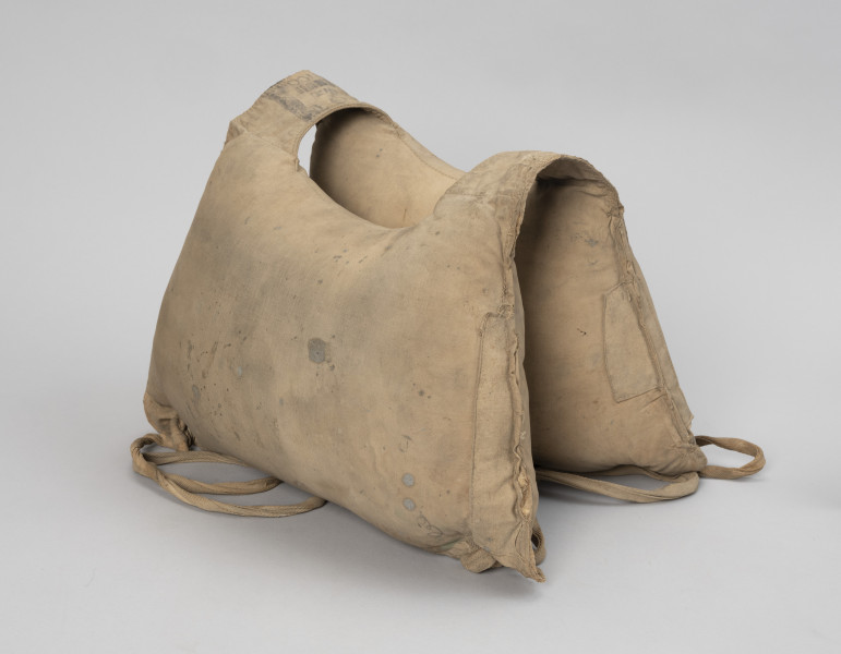 kamizelka ratunkowa - Ujęcie z boku; Kamizelka wykonana z dwóch pływaków płóciennych, przednim i tylnym, wypełnionych trawą morską, o kształtach zbliżonych do prostokąta lub trapezu (64 x 40 cm) z nieco wklęsłymi górnymi bokami. Górne narożniki pływaków połączone ze sobą pustymi komorami umożliwiającymi swobodne zwisanie dwóch pozostałych komór-pływaków, tworząc jednocześnie otwór na środku na głowę o średnicy 18 cm. Przy dolnych narożnikach kamizelki przyszyte przelotki z taśmy parcianej. W obydwu parach przelotek, wzdłuż dłuższych boków umieszczono taśmy parciane o długości 9 cm, zakończone stoperami powstałymi ze złożenia i przeszycia końcówek taśm, które zapobiegają wysunięciu się taśm z przelotek. Na jednym z dłuższych boków, przy szwach naszyte dodatkowe kawałki tkaniny wzmacniające naderwane szwy.