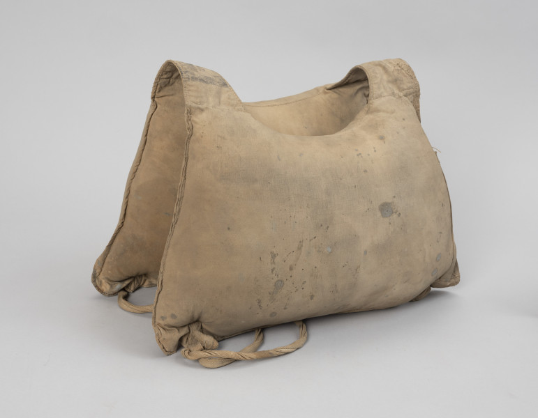 kamizelka ratunkowa - Ujęcie z tyłu; Kamizelka wykonana z dwóch pływaków płóciennych, przednim i tylnym, wypełnionych trawą morską, o kształtach zbliżonych do prostokąta lub trapezu (64 x 40 cm) z nieco wklęsłymi górnymi bokami. Górne narożniki pływaków połączone ze sobą pustymi komorami umożliwiającymi swobodne zwisanie dwóch pozostałych komór-pływaków, tworząc jednocześnie otwór na środku na głowę o średnicy 18 cm. Przy dolnych narożnikach kamizelki przyszyte przelotki z taśmy parcianej. W obydwu parach przelotek, wzdłuż dłuższych boków umieszczono taśmy parciane o długości 9 cm, zakończone stoperami powstałymi ze złożenia i przeszycia końcówek taśm, które zapobiegają wysunięciu się taśm z przelotek.