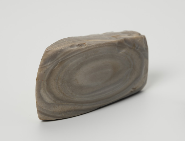 siekiera krzemienna - Ujęcie z góry; Siekiera czworościenna z grubym obuchem i łukowatym ostrzem. Ścianki szersze trapezowate, obuch czworoboczny lekko soczewkowaty. Krzemień pasiasty o szarych, jasnoszarych i beżowoszarych smugach z rudawymi przebarwieniami, na jednej z szerszych ścianek siekiery układającymi się koncentrycznie w owal. Powierzchnia szlifowana i gładzona.