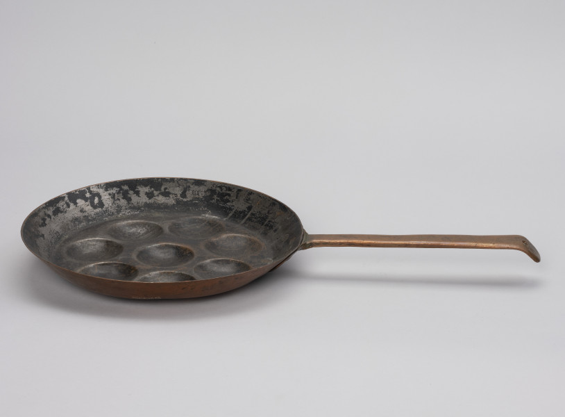 patelnia dołkownica - Ujęcie z boku; Patelnia wykonana z blachy miedzianej. W talerzu patelni 8 okrągłych, dość płytkich wgłębień. Kołnierz patelni wyskoki. Do kołnierza z prawej mocowana za pomocą 3 nitów długa, prosta rączka. Koniec rączki wygięty, lekko zaokrąglony z wyciętym okrągłym otworem służącym do powieszenia patelni na ścianie. Spodnia część patelni i rączka w dobrym stanie - miedź błyszcząca, powierzchnia wewnętrzna zdarta.