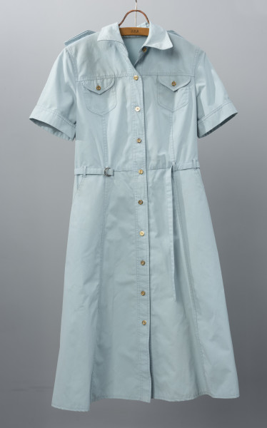 sukienka letnia z paskiem firmy DANA - Ujęcie z przodu; Jasnoniebieska sukienka z krótkimi rękawami i kołnierzykiem. Długość spódnicy za kolana. W talii cienki pasek z tego samego materiału co całość trzymany przez szlufki, zakończony niewielką, metalową sprzączką. Na wysokości klatki piersiowej dwie kieszenie, jedna po prawej, druga po lewej stronie, obie zapinane na metalowy guzik. Sukienka zapinana od góry do dołu na małe metalowe guziki. Materiał nieco sztywny.