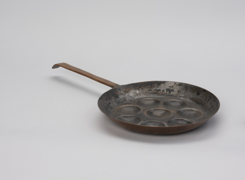 patelnia dołkownica - Ujęcie ze skosu; Patelnia wykonana z blachy miedzianej. W talerzu patelni 8 okrągłych, dość płytkich wgłębień. Kołnierz patelni wyskoki. Do kołnierza mocowana za pomocą 3 nitów długa, prosta rączka. Koniec rączki wygięty, lekko zaokrąglony. Spodnia część patelni i rączka w dobrym stanie - miedź błyszcząca, powierzchnia wewnętrzna zdarta.