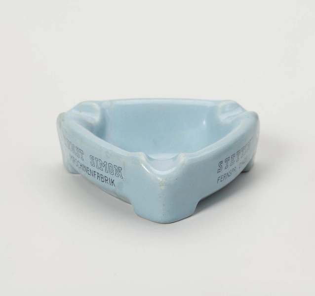 popielniczka reklamowa - Ujęcie z przodu; Popielniczka ceramiczna w kolorze szaroniebieskim o kształcie trójkąta równoramiennego z zaokrąglonymi narożnikami. Wywinięty na zewnątrz kołnierz tworzy boki naczynia, wydłużone na narożnikach w formę nóżek-podstawek. W górnej wypłaszczonej części kołnierza znajdują się zagłębienia służące do odłożenia papierosa. Na dwóch bocznych ściankach napisy reklamowe w 2 wierszach ciemnoniebieskimi literami.
