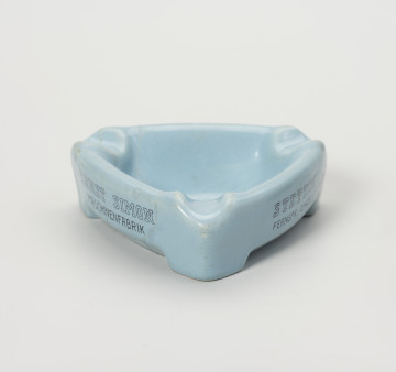 popielniczka reklamowa - Ujęcie z przodu; Popielniczka ceramiczna w kolorze szaroniebieskim o kształcie trójkąta równoramiennego z zaokrąglonymi narożnikami. Wywinięty na zewnątrz kołnierz tworzy boki naczynia, wydłużone na narożnikach w formę nóżek-podstawek. W górnej wypłaszczonej części kołnierza znajdują się zagłębienia służące do odłożenia papierosa. Na dwóch bocznych ściankach napisy reklamowe w 2 wierszach ciemnoniebieskimi literami.