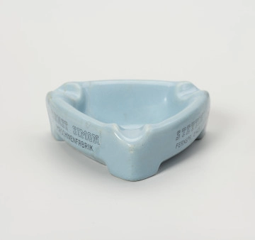 popielniczka reklamowa - Ujęcie z przodu; Popielniczka ceramiczna w kolorze szaroniebieskim o kształcie trójkąta równoramiennego z zaokrąglonymi narożnikami. Wywinięty na zewnątrz kołnierz tworzy boki naczynia, wydłużone na narożnikach w formę nóżek-podstawek. W górnej wypłaszczonej części kołnierza znajdują się zagłębienia służące do odłożenia papierosa. Na dwóch bocznych ściankach napisy reklamowe w 2 wierszach ciemnoniebieskimi literami.