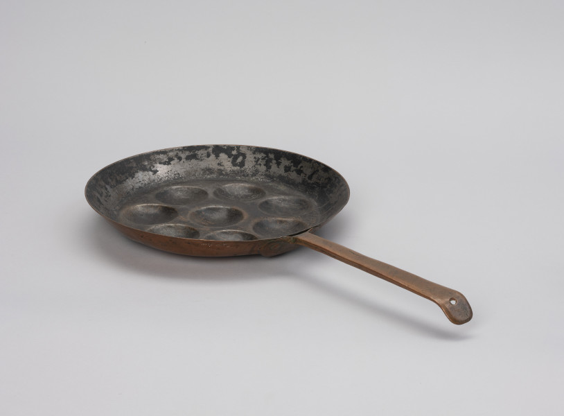 patelnia dołkownica - Ujęcie z tyłu; Patelnia wykonana z blachy miedzianej. W talerzu patelni 8 okrągłych, dość płytkich wgłębień. Kołnierz patelni wyskoki. Do kołnierza mocowana za pomocą 3 nitów długa, prosta rączka. Koniec rączki wygięty, lekko zaokrąglony z wyciętym okrągłym otworem służącym do powieszenia patelni na ścianie. Spodnia część patelni i rączka w dobrym stanie - miedź błyszcząca, powierzchnia wewnętrzna zdarta.