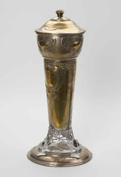 puchar klubu sportowego S.C. Blücher Stettin - ujęcie przodu ze skosu z lewej; Metalowe naczynie z pokrywą i podstawą, wydłużone w pionie. Podstawa złożona z okrągłej, lekko wysklepionej stopy i wspartej na niej ażurowej, wykonanej z cyny części wspierającej czarę. Dolna część czary wyższa, smukła, cylindryczna rozszerza się ku górze, górna wybrzuszona część jest wydzielona od dołu i od góry pierścieniami. Pokrywa okrągła, wysklepiona, z uchwytem w formie gałki ozdobionej rozetką, na wierzchołku. Powierzchnia pucharu zdobiona wypukłym ornamentem secesyjnym. Na stopie profilowanie wokół otoku, a w górnej części potrójna linia dekoracji z czterema falistymi załamaniami. Analogiczny ornament zdobi pokrywę. Część ażurowa ukształtowana z asymetrycznych zwojów miękko wyciętej taśmy, przechodzącej w cztery nóżki mocowane na śruby do stopy i tyle samo wsporników korpusu, z których dwa przykryte są ułożonymi w bukiet trzema wielopłatkowymi kwiatkami. Ze środkowego kwiatka wyprowadzona jest symetryczna dekoracja dolnej części czary, złożona z ornamentu roślinno-geometrycznego: pionowy trzon rozdwaja się sercowo w wić zakończoną kwiatkami, wyżej powtórnie otacza pole o kształcie medalionu, a następnie wygina się i prowadzi poziomo wzdłuż poszerzenia czary wicią z kwiatami i owocami. W centrum medalionu grawerowana tarcza z głową gryfa i napisem S.C. BLÜCHER STETTIN. W górnej części korpusu cztery symetryczne motywy ułożone w trójkątną formę z miękko wygiętych taśm; u dołu w obrębie pozbawionego dekoracji pola grawerowany napis: Meisterschaft 1900-10 .