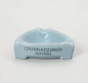 popielniczka reklamowa - Ujęcie z przodu; Popielniczka ceramiczna w kolorze szaroniebieskim o kształcie trójkąta równoramiennego z zaokrąglonymi narożnikami. Wywinięty na zewnątrz kołnierz tworzy boki naczynia, wydłużone na narożnikach w formę nóżek-podstawek. W górnej wypłaszczonej części kołnierza znajdują się zagłębienia służące do odłożenia papierosa. Na ściance napis reklamow w 2 wierszach ciemnoniebieskimi literami: CENTRALHEIZUNGEN | AUF ZUGE