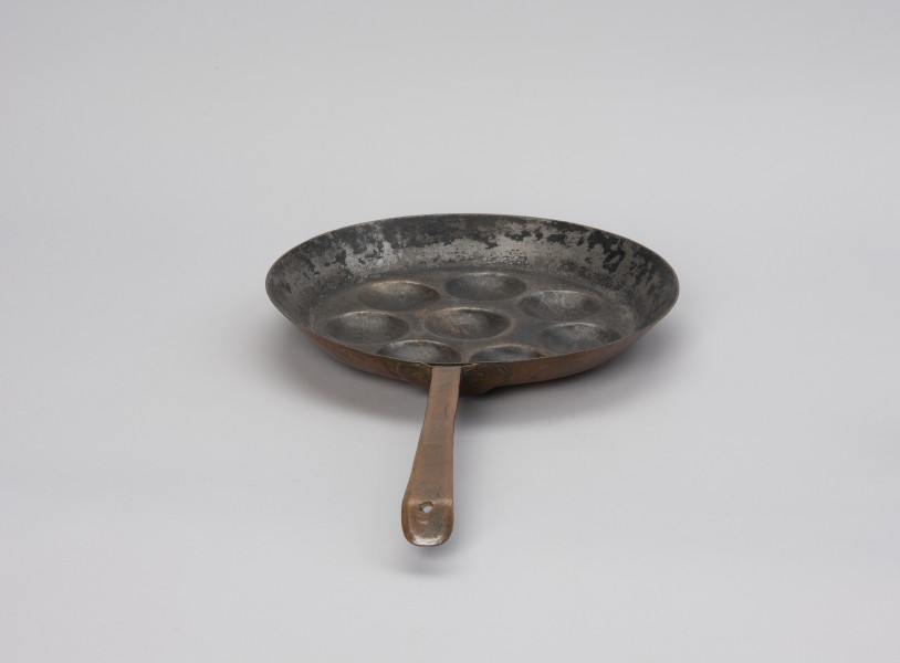 patelnia dołkownica - Ujęcie z tyłu; Patelnia wykonana z blachy miedzianej. W talerzu patelni 8 okrągłych, dość płytkich wgłębień. Kołnierz patelni wyskoki. Do kołnierza mocowana za pomocą 3 nitów długa, prosta rączka. Koniec rączki wygięty, lekko zaokrąglony z wyciętym okrągłym otworem służącym do powieszenia patelni na ścianie. Rączka w dobrym stanie - miedź błyszcząca, powierzchnia wewnętrzna zdarta.