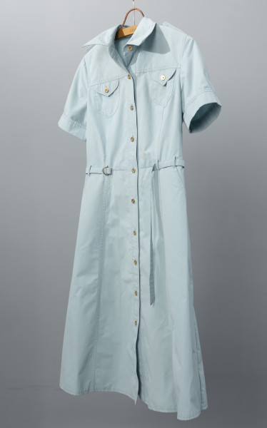 sukienka letnia z paskiem firmy DANA - Ujęcie ze skosu; Jasnoniebieska sukienka z krótkimi rękawami i kołnierzykiem. Długość spódnicy za kolana. W talii cienki pasek z tego samego materiału co całość trzymany przez szlufki, zakończony niewielką, metalową sprzączką. Na wysokości klatki piersiowej dwie kieszenie, jedna po prawej, druga po lewej stronie, obie zapinane na metalowy guzik. Sukienka zapinana od góry do dołu na małe metalowe guziki. Materiał nieco sztywny.
