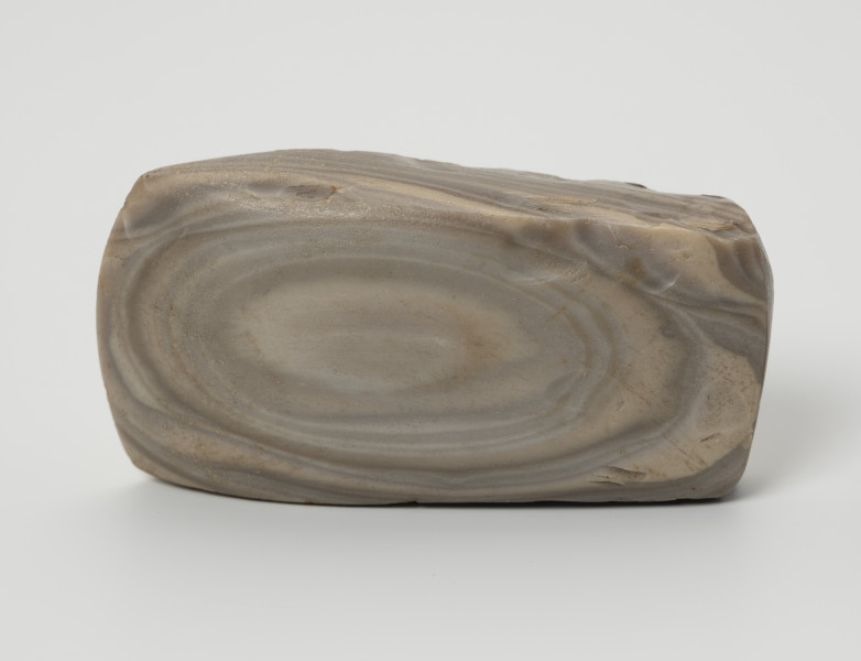 siekiera krzemienna - Ujęcie z góry; Siekiera czworościenna z grubym obuchem i łukowatym ostrzem. Krzemień pasiasty o szarych, jasnoszarych i beżowoszarych smugach z rudawymi przebarwieniami, na jednej z szerszych ścianek siekiery układającymi się koncentrycznie w owal. Powierzchnia szlifowana i gładzona.