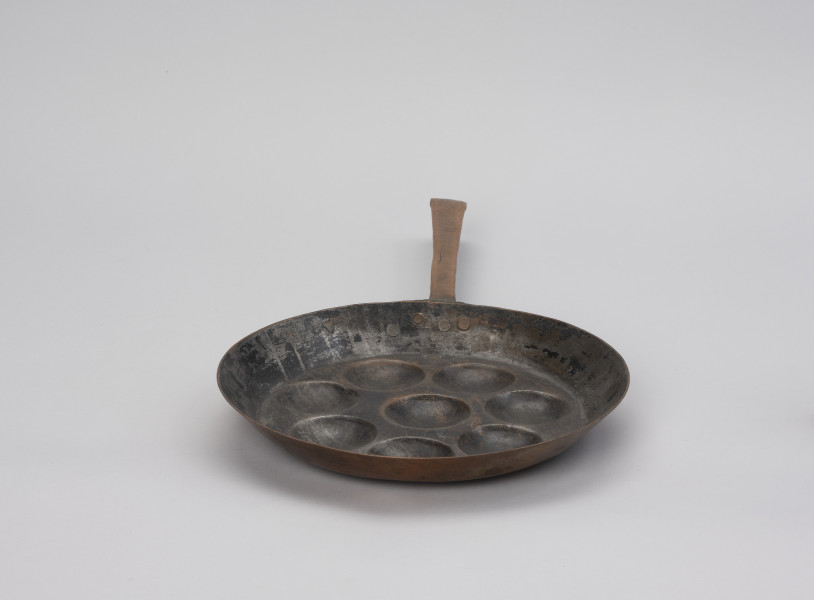 patelnia dołkownica - Ujęcie z tyłu; Patelnia wykonana z blachy miedzianej. W talerzu patelni 8 okrągłych, dość płytkich wgłębień. Kołnierz patelni wyskoki. Do kołnierza mocowana za pomocą 3 nitów długa, prosta rączka. Koniec rączki wygięty. Powierzchnia wewnętrzna zdarta.