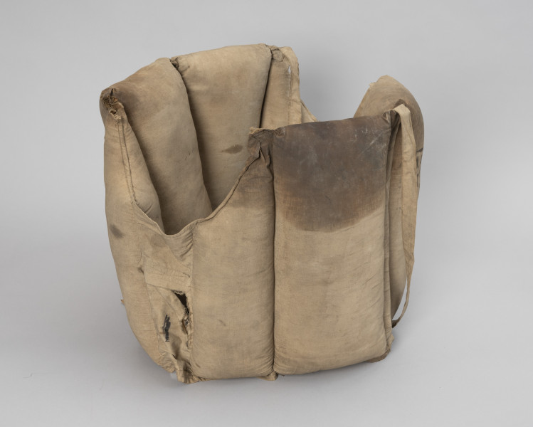 kamizelka ratunkowa trawiasta - Ujęcie z tyłu; Kamizelka wykonana z dwóch kawałków płótna beżowego koloru o kształtach zbliżonych do prostokąta ( 135x x 38 cm) złożona z ośmiu komór wypornościowych, cztery o wymiarach 17,5 x 38, cztery o wymiarach 10x38 z wyprofilowanym wycięciem na głowę, dodatkowo dwie komory puste o wymiarach 26x 8 cm umożliwiające swobodne zwisanie sąsiednich komór, pływaki wypełnione trawą morską. Krawędzie płócien zszyte ze sobą. Do dwóch mniejszych pływaków z wycięciem na głowę przyszyta parciana przelotka o wym. 46 x 2 cm. Jedna z pustych komór uszkodzona- naderwany materiał nad wszytym wzmocnieniem kieszeni. Całość nosi ślady eksploatacji z widocznymi tłustymi plamami.