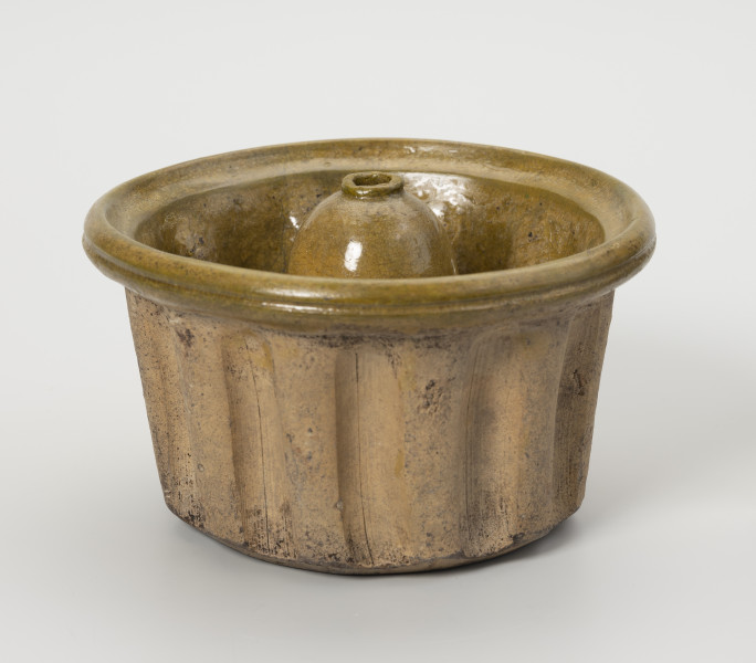 forma do wypieku babki - Ujęcie z przodu; Forma do wypieku babki o niewielkich rozmiarach i typowym dla tego rodzaju ciasta kształcie. Wysoki, prosty brzusiec regularnie karbowany i nieznacznie rozszerzający się ku górze przechodzi w wyraźnie wygięty na zewnątrz kołnierz o pogrubionej krawędzi. Centralna część dna wpuszczona do środka tworzy tak zwany komin (walec o kopulastym szczycie zakończonym małym otworkiem z zaznaczonym wylewem). Forma wykonana z gliny, wewnątrz jasnozielone szkliwo, zewnętrzna strona nie szkliwiona.