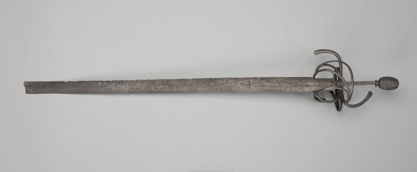 rapier - Ujęcie z przodu; Rapier o głowni prostej, obusiecznej, z ricassem. Na ricassie i krótkim odcinku głowni biegnie zaznaczone dwoma korytkami zbrocze. Sztych jest ułamany. Rękojeść ma krzyżowy jelec w esowatej formie i rozbudowany kosz do ochrony dłoni. Kosz składa się z trzech obłęków bocznych rozchodzących się ku ostrzu i osłaniających ricasso, po drugiej stronie są dwa obłęki boczne, z których górny jest większy, połączonych „oślą podkową” i dodatkowo dwoma obłękami. Trzon zwęża się ku końcowi, jego okładzina nie zachowała się. Głowica jest owalna, wydłużona z wyodrębnioną podstawą w formie pierścieni, ozdobiona jest wzdłużnie nacięciami i zakuta od góry.