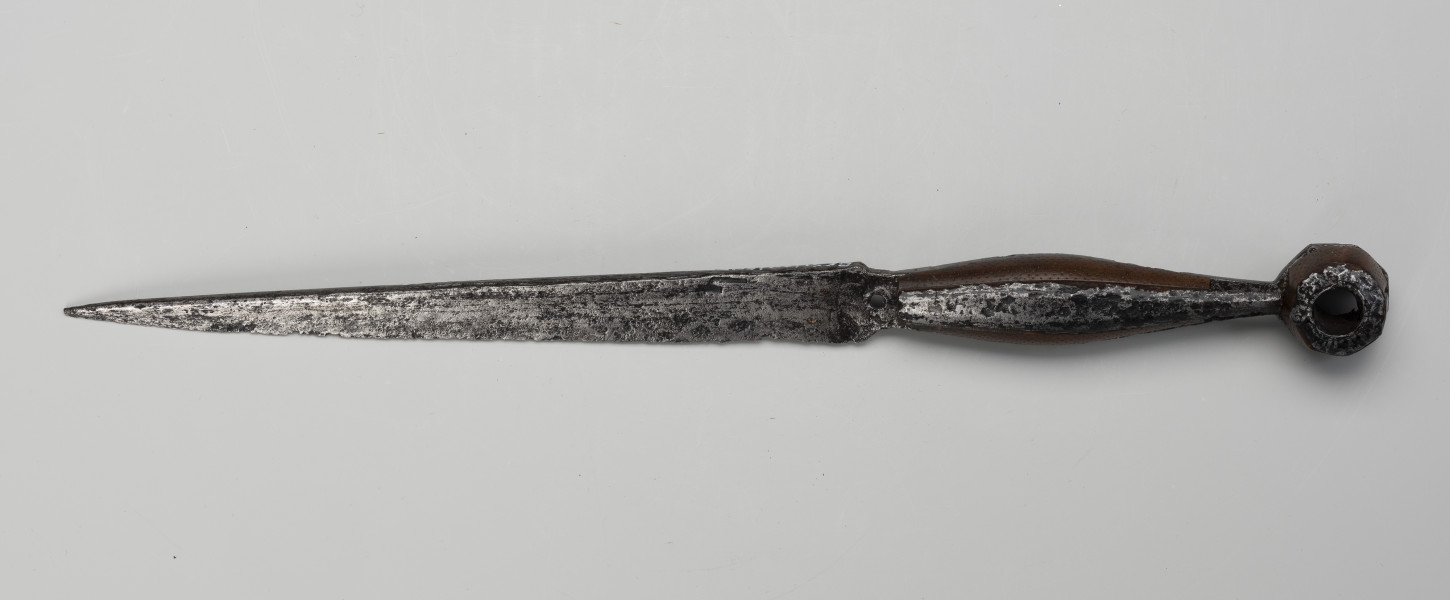 puginał - Ujęcie z przodu; Przybierający formę noża, jednosieczny puginał ma wąską głownię i rękojeść z pozostałościami okładzin rogowych na trzonie, które pierwotnie zamocowane były po jego drugiej stronie. Przymocowane zostały blaszanymi, gładkimi nakładkami, biegnącymi wzdłużnie pośrodku trzonka. Rogowe okładziny nabijane są srebrem, tworzącym punktowy wzór. Ośmiokątna, płaska głowica ze stali ma po środku okrągły otwór, przez który przechodzi tulejka, obłożona płytkami rogowymi oraz żelazna nakładka z rytymi motywami płatków kwiatów.