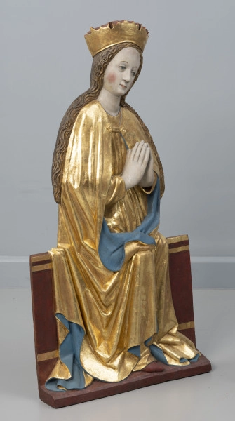 dwufigurowa grupa płaskorzeźb ołtarzowych - Ujęcie przodu ze skosu z lewej; Płaskorzeźbiona figura przedstawia na szerokiej ławie tronuje Maria. Postać ubrana jest w długą tunikę przewiązaną w pasie i płaszcz spięty klamrą powyżej piersi. Miękka, lejąca się ku podstawie rzeźby złocista tkanina, szata połyskuje efektownie marszczeniami o łagodnych grzbietach. Na głowie wysoka złocona korona o gładkiej obręczy, zwieńczona zygzakiem. Maria lekko przechyla głowę wlewo, dłonie trzymając w geście modlitewnym przy piersi. Jej młodzieńczą twarz o wysokim czole i łagodnych rysach otaczają pasma długich falujących, złocistych włosów.  Długie włosy opadają na ramiona. Tył rzeźby opracowany płasko. Fleurony wieńczące obręcze koron nie zachowały się. Złocenia płaskorzeźby częściowo rekonstruowane.