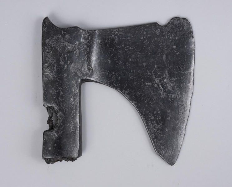 żeleźce topora, oksza typu X (Głosek) - Ujęcie z tyłu; Oksza w typie X (według typologii Mariana Głoska), o górnej krawędzi wznoszącej się lekko ku górze, dolnej opadającej łukiem. Broda lekko ścięta pod skosem, szyja szeroka. Ostrze jest asymetryczne i lekko wygięte. Osada jest długa, zakuta od góry, z płynnie wyprofilowanym zgrubieniem w górnej krawędzi przechodzącym w wystający kolec. Osada ma ubytki metalu spowodowane korozją. Wewnątrz osady zachowały się drewniane pozostałości trzonka.