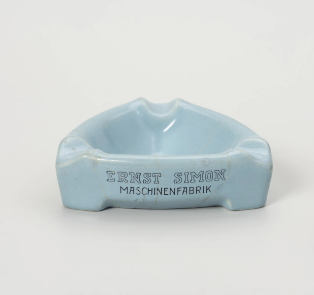 popielniczka reklamowa - Ujęcie z przodu; Popielniczka ceramiczna w kolorze szaroniebieskim o kształcie trójkąta równoramiennego z zaokrąglonymi narożnikami. Wywinięty na zewnątrz kołnierz tworzy boki naczynia, wydłużone na narożnikach w formę nóżek-podstawek. W górnej wypłaszczonej części kołnierza znajdują się zagłębienia służące do odłożenia papierosa. Na ściance napis reklamowy w 2 wierszach ciemnoniebieskimi literami: ERNST SIMON | MASCHINENFABRIK