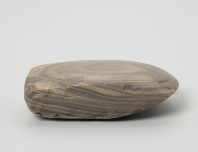 siekiera krzemienna - Ujęcie z boku; Siekiera czworościenna z grubym obuchem i łukowatym ostrzem. Ścianki szersze trapezowate, obuch czworoboczny lekko soczewkowaty. Krzemień pasiasty o szarych, jasnoszarych i beżowoszarych smugach z rudawymi przebarwieniami, na jednej z szerszych ścianek siekiery układającymi się koncentrycznie w owal. Powierzchnia szlifowana i gładzona.