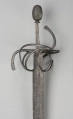 rapier - Ujęcie 3/4; Rapier o głowni prostej, obusiecznej, z ricassem. Na ricassie i krótkim odcinku głowni biegnie zaznaczone dwoma korytkami zbrocze. Rękojeść ma krzyżowy jelec w esowatej formie i rozbudowany kosz do ochrony dłoni. Kosz składa się z trzech obłęków bocznych rozchodzących się ku ostrzu i osłaniających ricasso, po drugiej stronie są dwa obłęki boczne, z których górny jest większy, połączonych „oślą podkową” i dodatkowo dwoma obłękami. Głowica jest owalna, wydłużona z wyodrębnioną podstawą w formie pierścieni, ozdobiona jest wzdłużnie nacięciami i zakuta od góry.