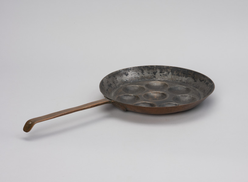 patelnia dołkownica - Ujęcie ze skosu; Patelnia wykonana z blachy miedzianej. W talerzu patelni 8 okrągłych, dość płytkich wgłębień. Kołnierz patelni wyskoki. Do kołnierza mocowana za pomocą 3 nitów długa, prosta rączka. Koniec rączki wygięty, lekko zaokrąglony z wyciętym okrągłym otworem służącym do powieszenia patelni na ścianie. Spodnia część patelni i rączka w dobrym stanie - miedź błyszcząca, powierzchnia wewnętrzna zdarta.