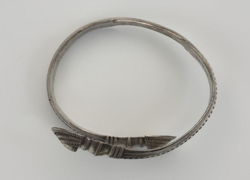 bransoleta żmijowata typu II wg Blumego - Ujęcie z góry; Bransoleta żmijowata typu II wg Ericha Blumego wykonana z srebrnej taśmy zgiętej w okręg, której zakończenia przypominają główki żmij. Maksymalna szerokość kabłąka to 14 mm i szerokości zakończeń 22 mm. Zakończenia kabłąka ornamentowane są karbowaniem i zwielokrotnionymi pasmami pseudofiligranu, cyzelowanymi w poprzek, a na samych łukowatych główkach zakończeń koncentrycznie wzdłuż krawędzi.
