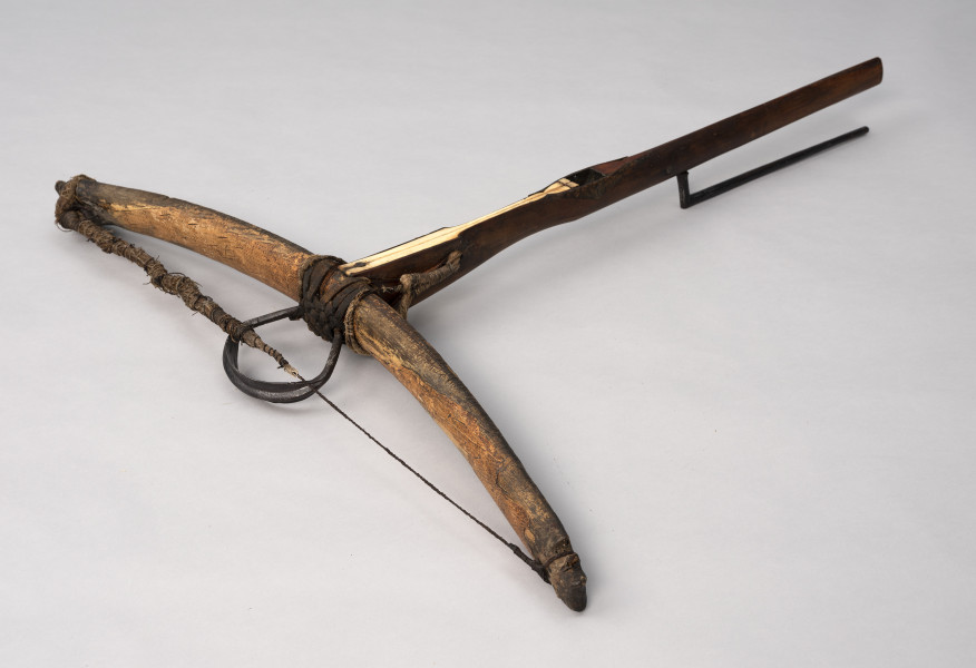 kusza - Ujęcie ze skosu; Kusza średniowieczna z odgiętym łęczyskiem, drewnianym łożem z kościaną nakładką i prostą konstrukcją dźwigni spustowej. Łoże jest proste, zwęża się ku końcowi kolby. Od góry na łożu zamocowana jest kościana płytka z wgłębieniem na prowadnicę bełta przytwierdzona w najszerszej części gwoździem. Zwęża się ku otworowi na orzech, a przed nim znowu się rozszerza. Widoczna część spustu jest prosta, następnie wygina się pod kątem 90 stopni.  Łuczysko jest mocowane do frontu łoża wspomnianym oplotem. Jest wygięte na zewnątrz i pokryte korą drzewa brzozowego. Na lewym końcu łuku widoczne jest wgłębienie do mocowania cięciwy. Zachowana cięciwa jest urwana składa się ze splotu wielu cienkich nici. Pośrodku łuku przywiązane jest rzemieniem żelazne strzemię, zdobione po bokach nacięciami.