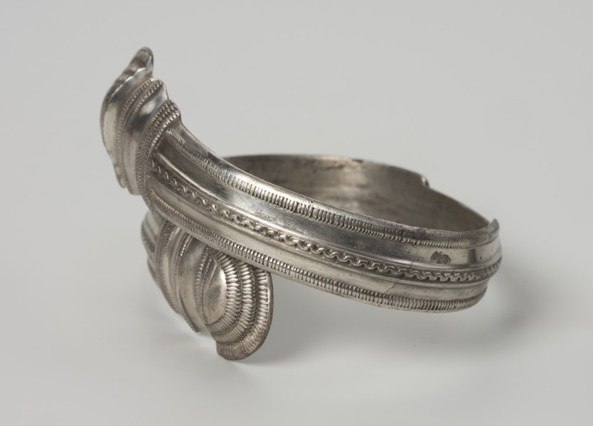 bransoleta żmijowata typu II wg Blumego - Ujęcie z boku; Bransoleta żmijowata typu II wg Ericha Blumego wykonana z srebrnej taśmy zgiętej w okręg, której zakończenia przypominają główki żmij. Maksymalna szerokość kabłąka to 14 mm i szerokości zakończeń 22 mm. Powierzchnia kabłąka zdobiona trzema wzdłużnymi pasmami ornamentu: pseudofiligranem przy krawędziach zewnętrznych i wężykiem ujętym w dwa wąskie żeberka pośrodku. Zakończenia kabłąka ornamentowane są karbowaniem i zwielokrotnionymi pasmami pseudofiligranu, cyzelowanymi w poprzek, a na samych łukowatych główkach zakończeń koncentrycznie wzdłuż krawędzi.