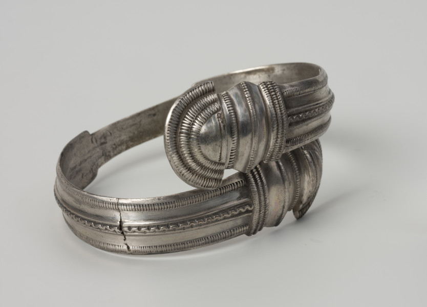 bransoleta żmijowata typu II wg Blumego - Ujęcie z przodu; Bransoleta żmijowata typu II wg Ericha Blumego wykonana z srebrnej taśmy zgiętej w okręg, której zakończenia przypominają główki żmij. Maksymalna szerokość kabłąka to 14 mm i szerokości zakończeń 22 mm. Powierzchnia kabłąka zdobiona trzema wzdłużnymi pasmami ornamentu: pseudofiligranem przy krawędziach zewnętrznych i wężykiem ujętym w dwa wąskie żeberka pośrodku. Zakończenia kabłąka ornamentowane są karbowaniem i zwielokrotnionymi pasmami pseudofiligranu, cyzelowanymi w poprzek, a na samych łukowatych główkach zakończeń koncentrycznie wzdłuż krawędzi.