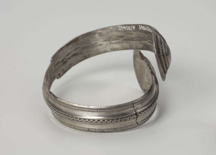 bransoleta żmijowata typu II wg Blumego - Ujęcie z boku; Bransoleta żmijowata typu II wg Ericha Blumego wykonana z srebrnej taśmy zgiętej w okręg, której zakończenia przypominają główki żmij. Maksymalna szerokość kabłąka to 14 mm i szerokości zakończeń 22 mm. Powierzchnia kabłąka zdobiona trzema wzdłużnymi pasmami ornamentu: pseudofiligranem przy krawędziach zewnętrznych i wężykiem ujętym w dwa wąskie żeberka pośrodku.