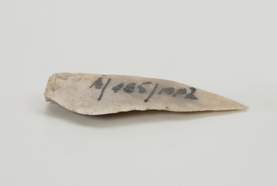 półwytwór liściaka - Ujęcie z boku; Krótki wiór w kształcie wydłużonego trójkąta, odbity miękkim tłukiem (prawdopodobnie z poroża) od większej bryły. Brakuje retuszu, który formowałby krótki trzonek. Ze względu na jego rozmiary prawdopodobnie miał służyć jako grot włóczni.