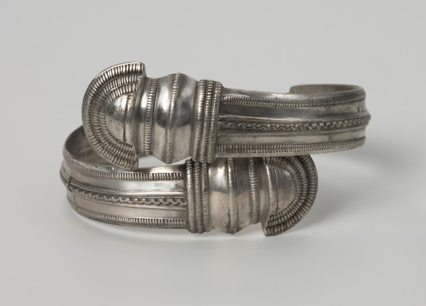 bransoleta żmijowata typu II wg Blumego - Ujęcie z przodu; Bransoleta żmijowata typu II wg Ericha Blumego wykonana z srebrnej taśmy zgiętej w okręg, której zakończenia przypominają główki żmij. Maksymalna szerokość kabłąka to 14 mm i szerokości zakończeń 22 mm. Powierzchnia kabłąka zdobiona trzema wzdłużnymi pasmami ornamentu: pseudofiligranem przy krawędziach zewnętrznych i wężykiem ujętym w dwa wąskie żeberka pośrodku. Zakończenia kabłąka ornamentowane są karbowaniem i zwielokrotnionymi pasmami pseudofiligranu, cyzelowanymi w poprzek, a na samych łukowatych główkach zakończeń koncentrycznie wzdłuż krawędzi.