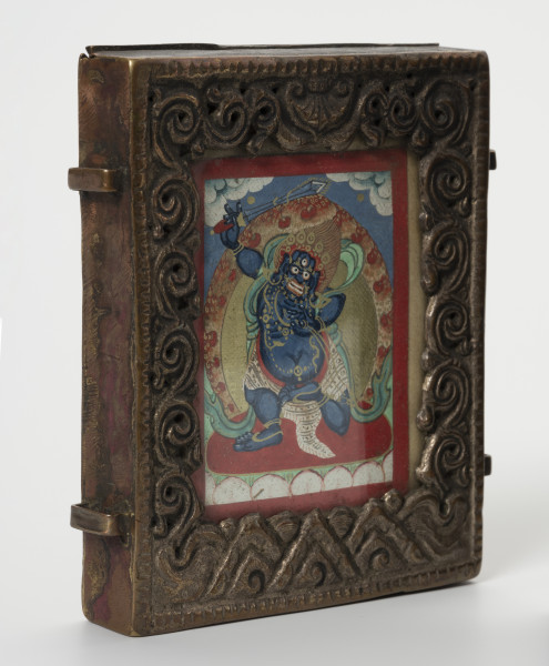 relikwiarz gau z miniaturą sakralną tsakli - ujęcie przodu ze skosu z lewej; Miedziany relikwiarz w formie małej, wąskiej, przeszklonej i bogato zdobionej ramki wypełnionej obrazem Wadżrapani (sanskr. Vajrapāṇi, tyb. Cziano Dordże). Postać zaprezentowana w  formie gniewnej, najprawdopodobniej przedstawia Wadżrapani jako strażnika Dharmy (sanskr. dharamapāla) – Wadżrapani-Acharya. Ukazany jako tęgi i silny, z potężnym ciemnoniebieskim ciałem, wystającym brzuchem, grubymi, krótkimi kończynami i wściekłym wyrazem twarzy. W odmianie ikonograficznej z jedną głową, trzecim okiem, nastroszonymi włosami, pięcioramienną koroną bodhisattwy, naszyjnikami z węży oraz pasem z tygrysiej skóry. Otoczony płomieniami trzyma miecz i lasso (sanskr. pāśa). Stoi na lotosie – symbolu czystości i poznania, na tle nieba i chmur. Obrazek zakończony czerwoną bordiurą. Ramka ozdobiona prostymi nacięciami przy zewnętrznych krawędziach oraz misternie wykonanymi wzorami przy wewnętrznych: usytuowany po bokach wzór grawerowanej wici roślinnej, centralnie, w górnej części znajduje się przedstawienie przypominające baldachim (parasol buddyjski), natomiast zlokalizowane w dolnej części trójkątne motywy być może przedstawiają muszle - motyw konchy. Przy bocznych powierzchniach dwa prostokątne mocowania - ucha na pasek do noszenia relikwiarza, ułożone symetrycznie. Widoczne ślady użytkowania, liczne przetarcia, odbarwienia.