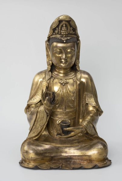 Bodhisattwa Guanyin - ujęcie z przodu; Duża, wykonana ze złoconego brązu figura przedstawia siedzącą w pozie medytacyjnej kobietę Bodhisattwę Guanyin (chin. 观音, sanskr. Awalokiteśwara, tyb. Czenrezig). Postać ma spuszczone, przymknięte oczy, symetryczną, pełną twarz o pogodnym i spokojnym wyrazie oraz gładko zaczesane włosy, schowane pod diademem i zaciągniętą na głowę tkaniną. Ubrana jest w długie, warstwowe, jedwabne szaty królewskie oraz wyszukaną, ciężką biżuterię książęcą: diadem, w którego centrum znajduje się niebiański Budda Amitabha (sanskr. Amitābha); pary dużych, okrągłych kolczyków ze spiczastymi zawieszkami oraz grubego naszyjnika z klejnotem, od którego odchodzi siedem pasów korali z wisiorkami. Jej prawa dłoń uniesiona na wysokości klatki piersiowej ułożona jest pionowo, jej środkowy palec i kciuk tworzą okrąg trzymając niezidentyfikowany, haczykowaty przedmiot. Poniżej znajduje się ustawiona poziomo lewa dłoń, która ściska niewielką wazę.