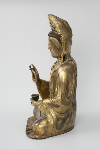 Bodhisattwa Guanyin - ujęcie z drugiego boku; Duża, wykonana ze złoconego brązu figura przedstawia siedzącą w pozie medytacyjnej kobietę Bodhisattwę Guanyin (chin. 观音, sanskr. Awalokiteśwara, tyb. Czenrezig). Postać ma spuszczone, przymknięte oczy, symetryczną, pełną twarz o pogodnym i spokojnym wyrazie oraz gładko zaczesane włosy i zaciągniętą na głowę tkaniną. Ubrana jest w długie, warstwowe, jedwabne szaty królewskie, pary dużych, okrągłych kolczyków ze spiczastymi zawieszkami. Jej prawa dłoń uniesiona na wysokości klatki piersiowej ułożona jest pionowo. Poniżej znajduje się ustawiona poziomo lewa dłoń, która ściska niewielką wazę.