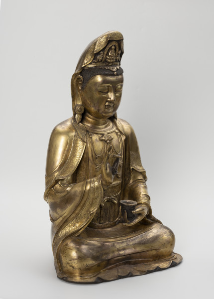 Bodhisattwa Guanyin - ujęcie przodu ze skosu z lewej; Duża, wykonana ze złoconego brązu figura przedstawia siedzącą w pozie medytacyjnej kobietę Bodhisattwę Guanyin (chin. 观音, sanskr. Awalokiteśwara, tyb. Czenrezig). Postać ma spuszczone, przymknięte oczy, symetryczną, pełną twarz o pogodnym i spokojnym wyrazie oraz gładko zaczesane włosy, schowane pod diademem i zaciągniętą na głowę tkaniną. Ubrana jest w długie, warstwowe, jedwabne szaty królewskie oraz wyszukaną, ciężką biżuterię książęcą: diadem, w którego centrum znajduje się niebiański Budda Amitabha (sanskr. Amitābha); pary dużych, okrągłych kolczyków ze spiczastymi zawieszkami oraz grubego naszyjnika z klejnotem, od którego odchodzi siedem pasów korali z wisiorkami. Jej prawa dłoń uniesiona na wysokości klatki piersiowej ułożona jest pionowo, jej środkowy palec i kciuk tworzą okrąg trzymając niezidentyfikowany, haczykowaty przedmiot. Poniżej znajduje się ustawiona poziomo lewa dłoń, która ściska niewielką wazę.